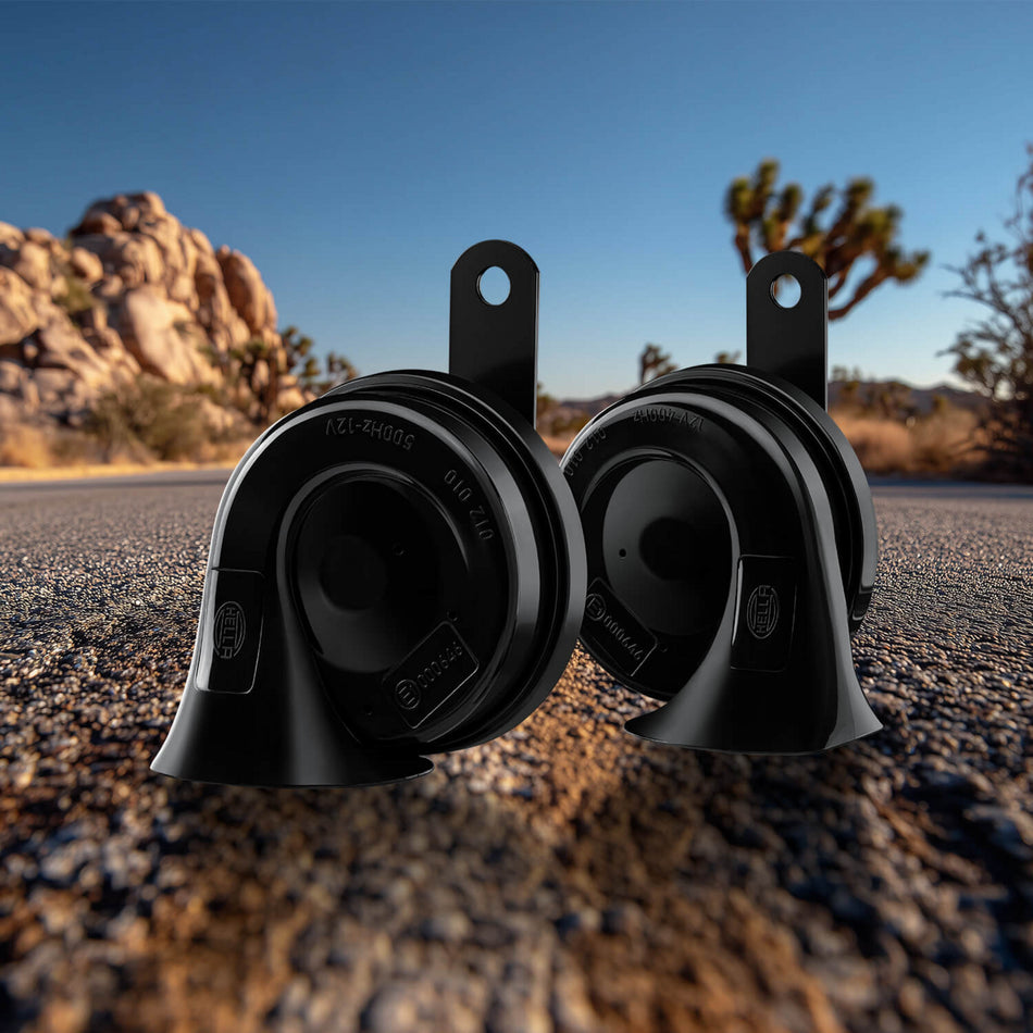 Two black HELLA TE26 OE Connection car horns with mounting brackets on an asphalt road.