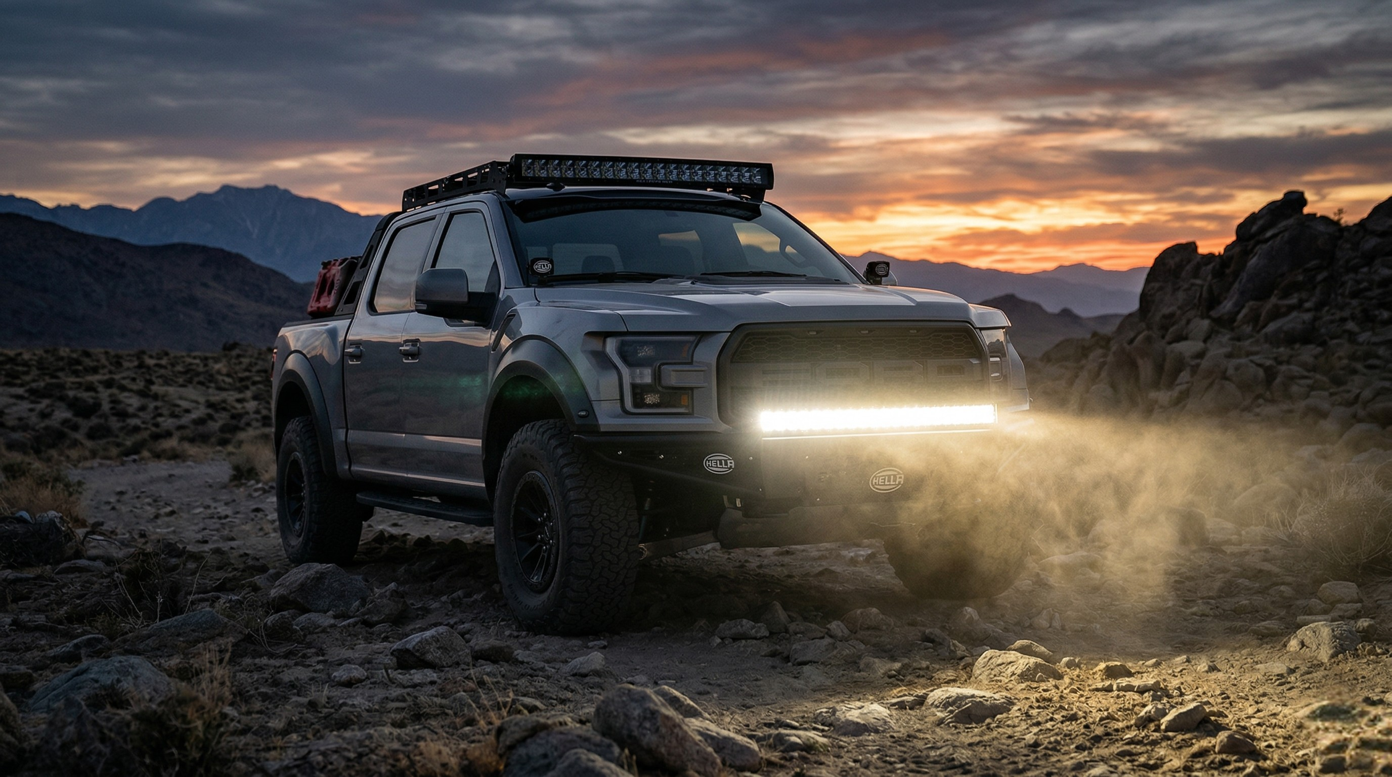 Ford F-150 Raptor off-road truck with illuminated HELLA Black Magic LED light bars, on a rocky desert trail during a dramatic sunset.